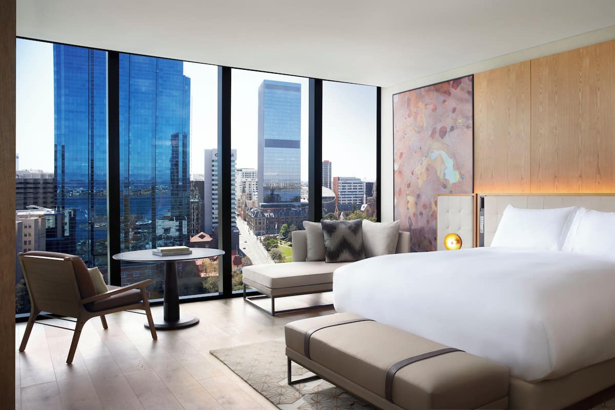Una habitación de hotel moderna con una cama grande, una mesita, una silla y vistas a la ciudad a través de amplios ventanales.