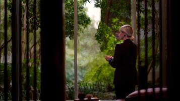 A person standing by a window with a view of greenery.