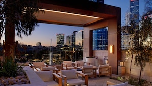 An outdoor seating area with multiple chairs and tables, a potted plant, and a view of the city skyline at dusk.