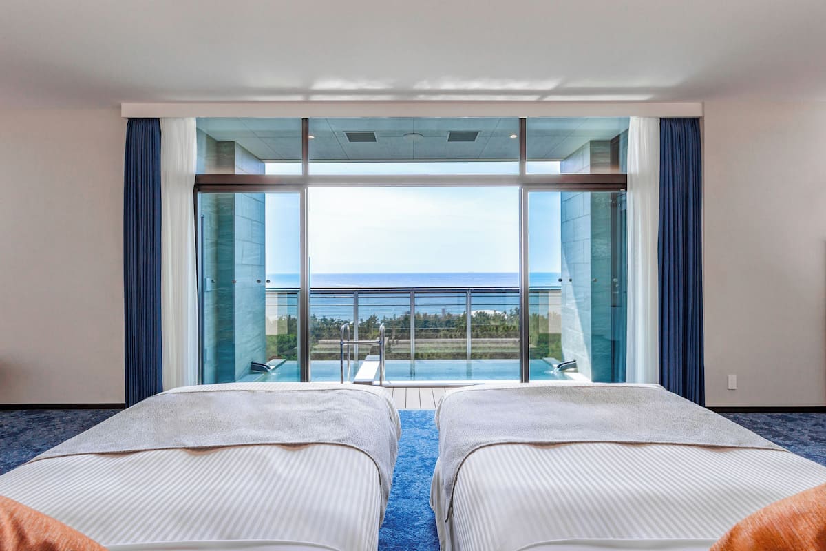 Habitación de hotel con dos camas, un ventanal con vistas al océano y un balcón con piscina.