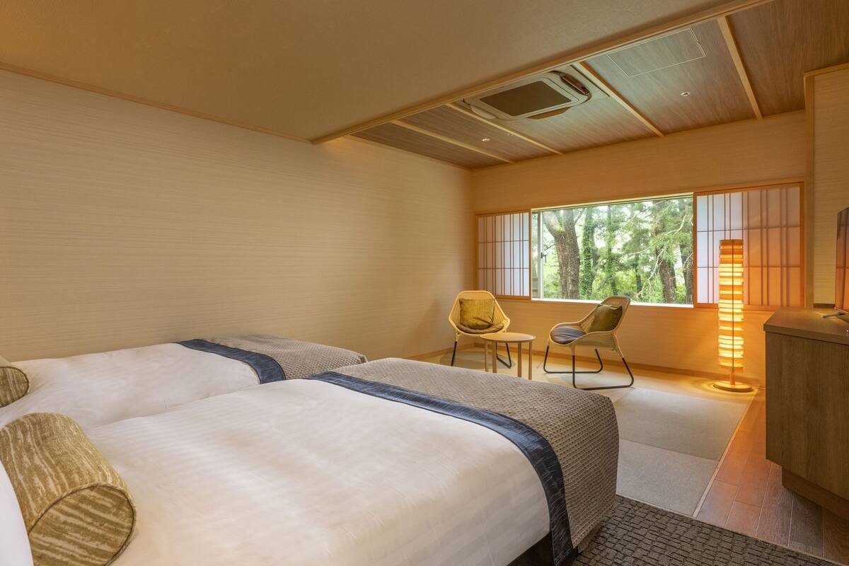 Habitación de hotel con dos camas, una mesita pequeña, dos sillas, un escritorio y una ventana con vista a árboles.