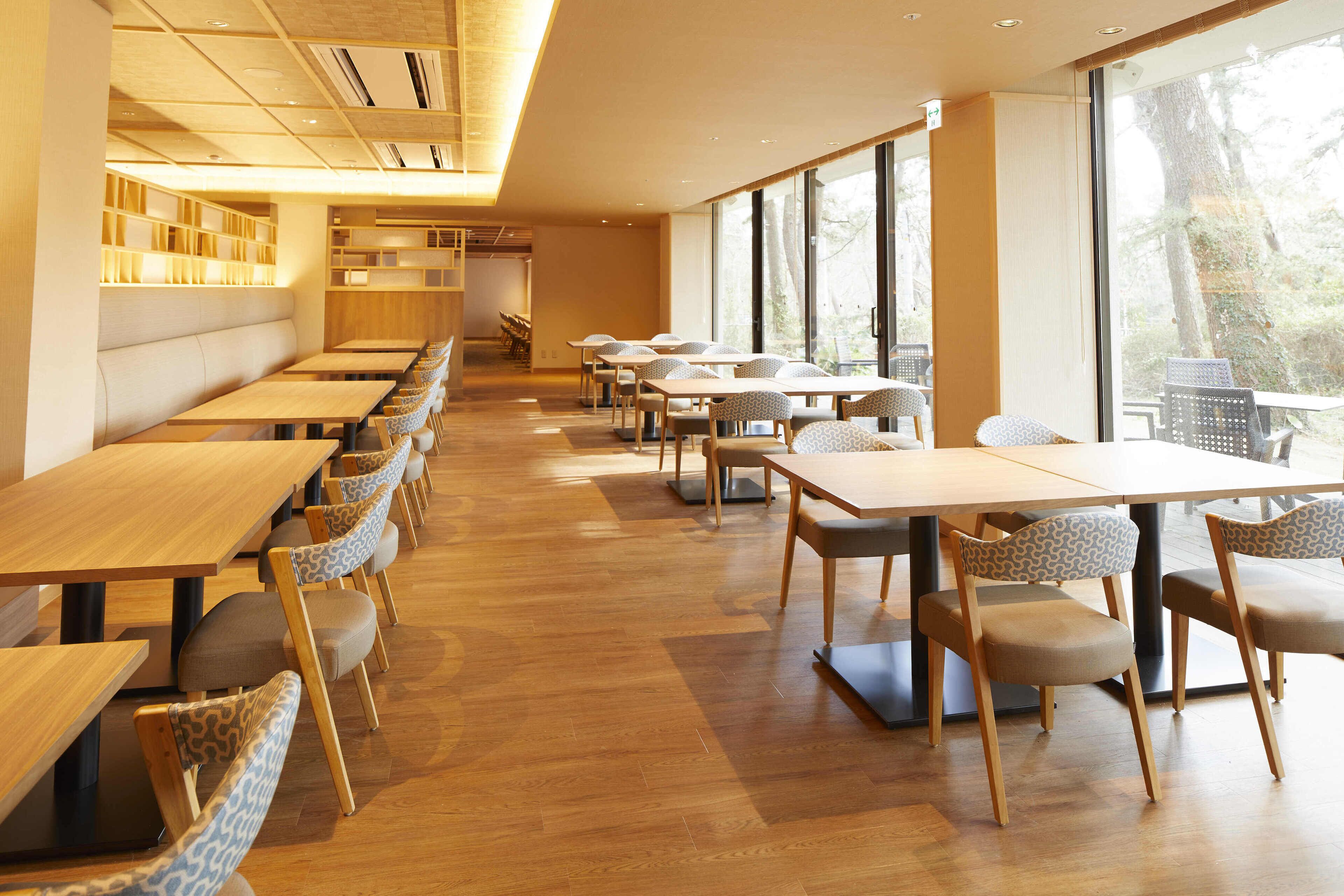 A modern restaurant with wooden tables and chairs, large windows, and a bright interior.