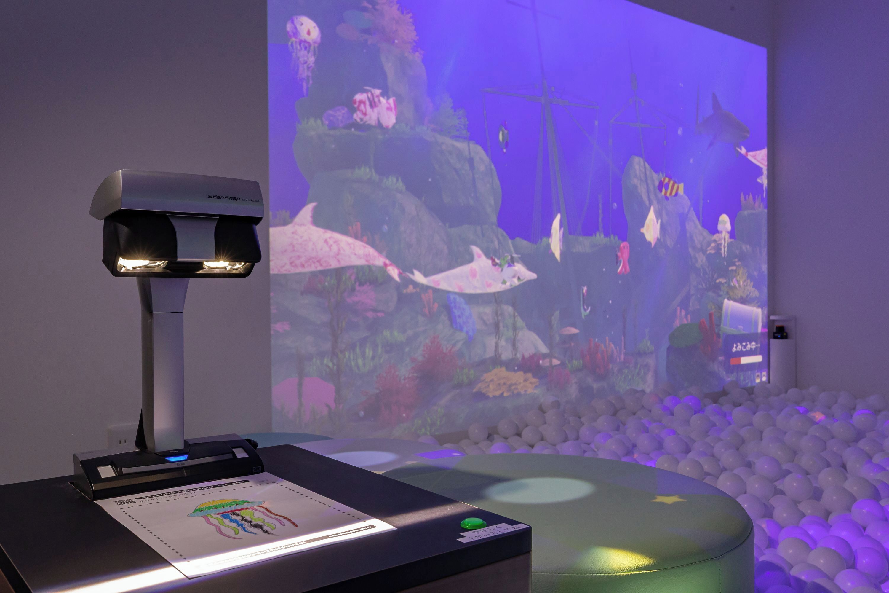 A room with a projection screen displaying an underwater scene, a desk with a scanner, and a ball pit.