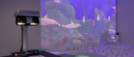 A room with a projection screen displaying an underwater scene, a desk with a scanner, and a ball pit.