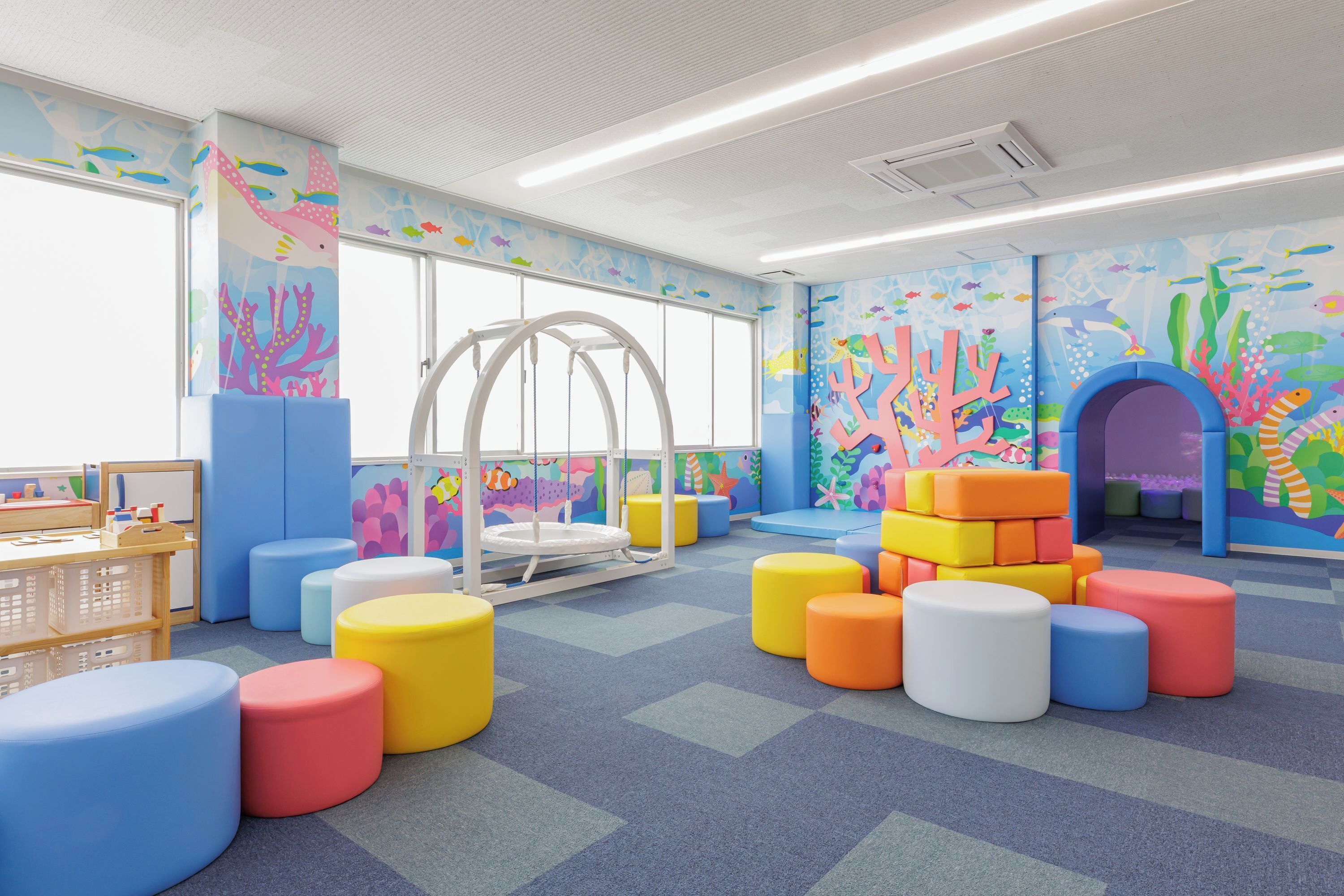 Zona infantil con taburetes de colores, una estructura de juegos y decoración mural con motivos marinos.