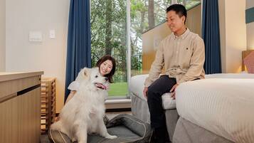 A hotel room with a bed, a pet bed, a wooden desk, and a view of greenery outside.