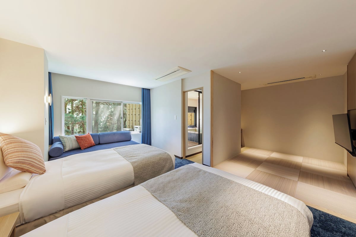 Una habitación de hotel moderna con dos camas, un ventanal con vistas a árboles, un sofá y un televisor de pantalla plana.