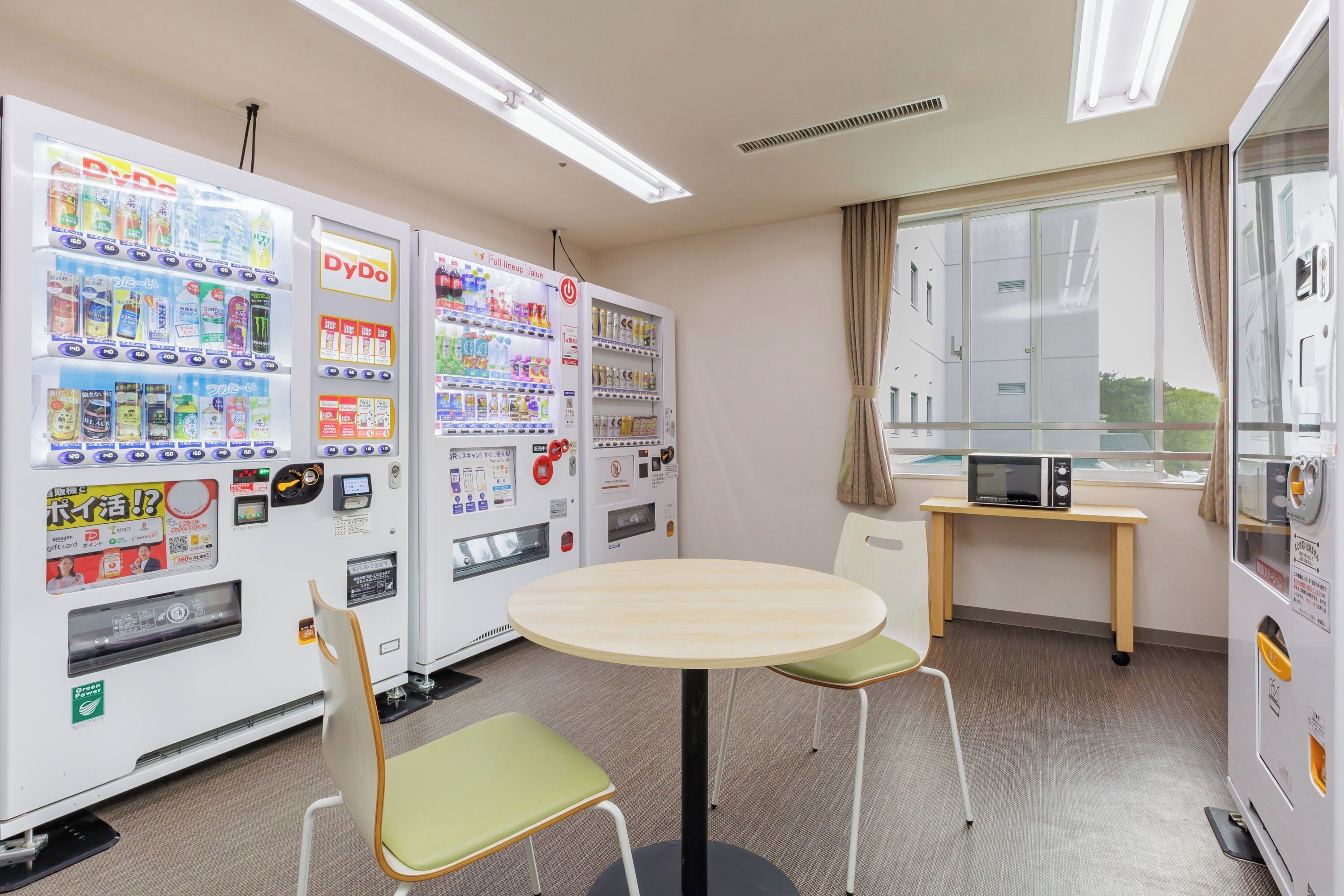 Una habitación con una mesa redonda, dos sillas, máquinas expendedoras, un microondas y una ventana con cortinas.