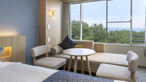 Habitación de hotel con cama, sofá, una mesita y un amplio ventanal que ofrece vistas a árboles y al mar.