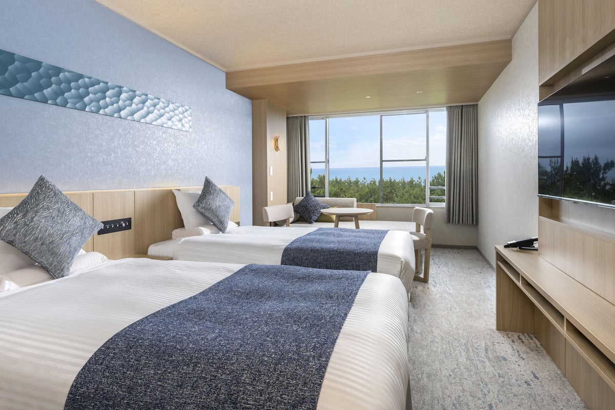 Habitación de hotel con dos camas, un sofá, un televisor y un ventanal con vista al mar.