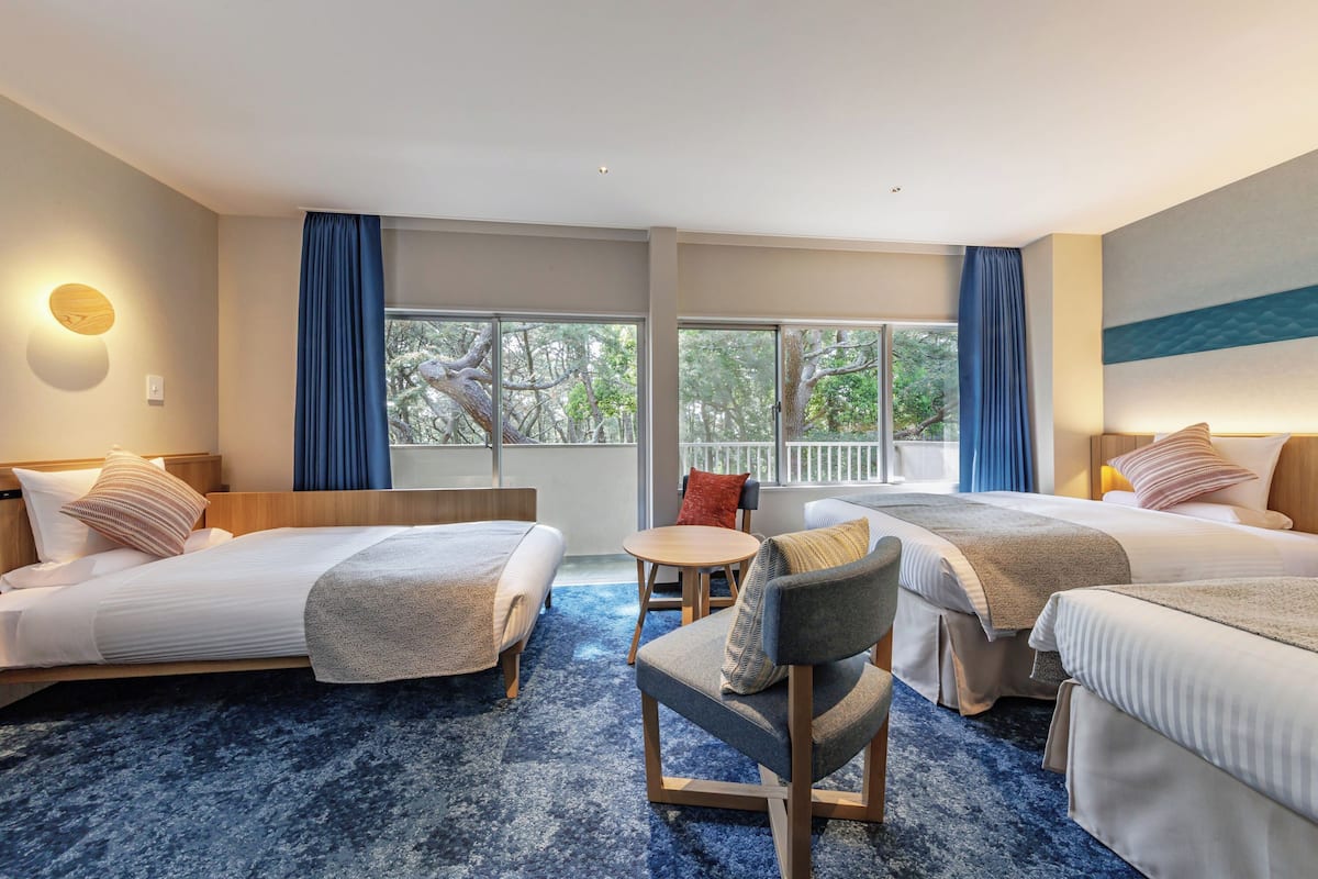 Habitación de hotel con dos camas, una mesita redonda pequeña y una silla. Cuenta con ventanas grandes con cortinas, vistas a árboles desde el exterior y un cuadro en la pared.