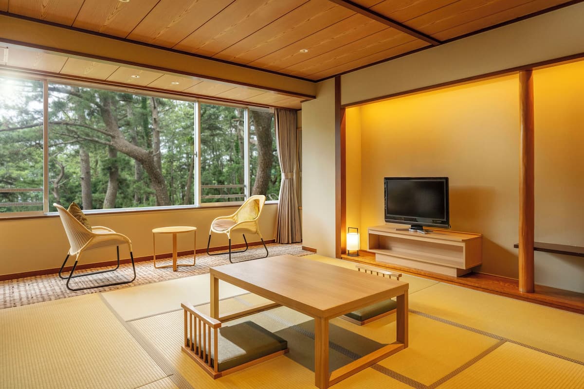 Una habitación japonesa tradicional con suelo de tatami, una mesa de madera baja, dos sillas, un televisor y una puerta corredera que da a vistas de árboles.