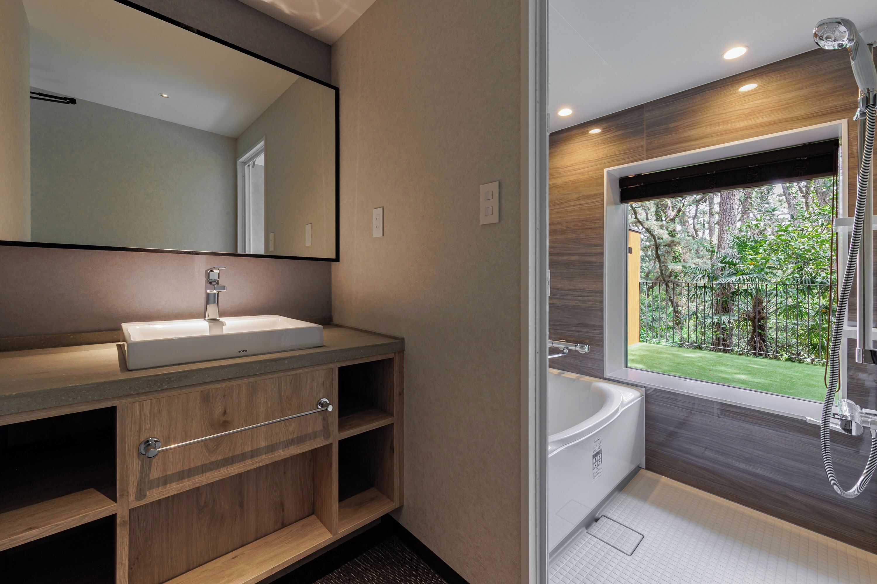 Een moderne badkamer met een grote spiegel, een houten wastafelmeubel, een witte wastafel en een bad met uitzicht op een tuin.