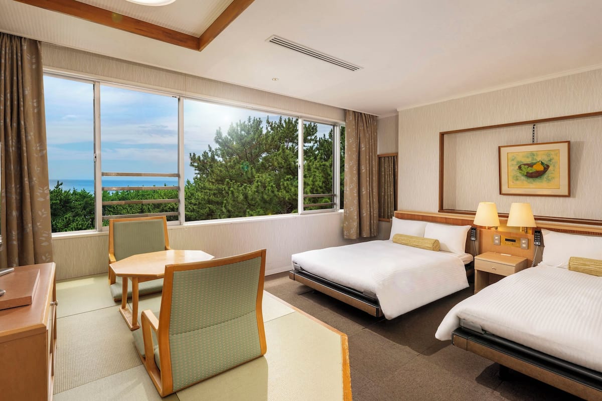 Habitación de hotel con un ventanal grande que ofrece vistas al mar, una cama, un escritorio con silla y una mesita de noche con lámparas.