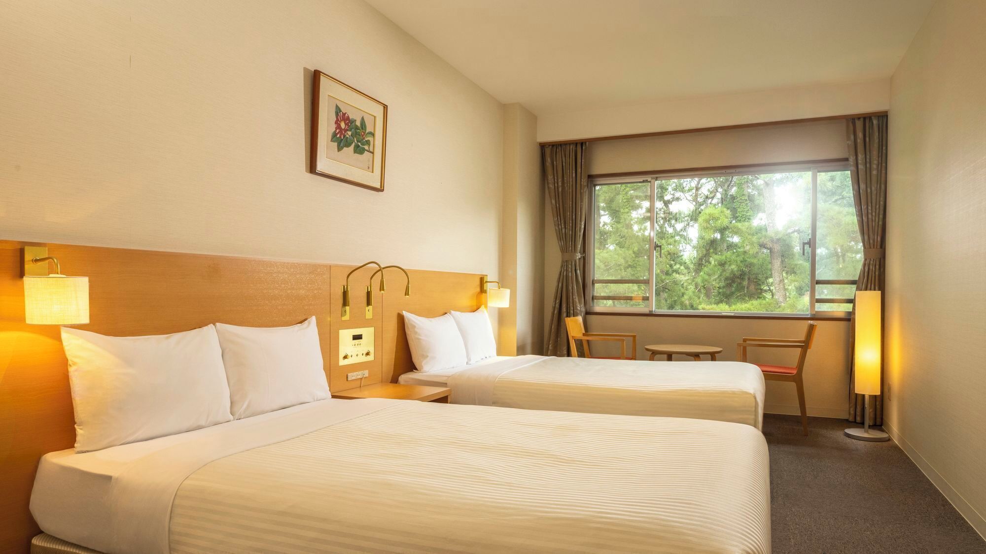 A hotel room with two beds, a small table, a chair, a window with curtains, and a wall-mounted lamp.