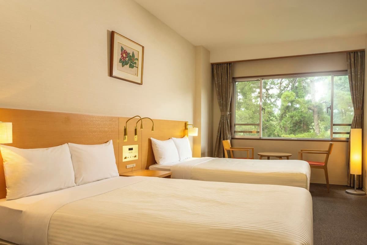 Habitación de hotel con dos camas, una mesita, una silla, una ventana con cortinas y una lámpara de pared.