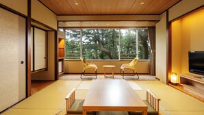 Una habitación japonesa tradicional con suelo de tatami, una mesa de madera baja, dos sillas, un televisor y un ventanal con vistas a árboles.
