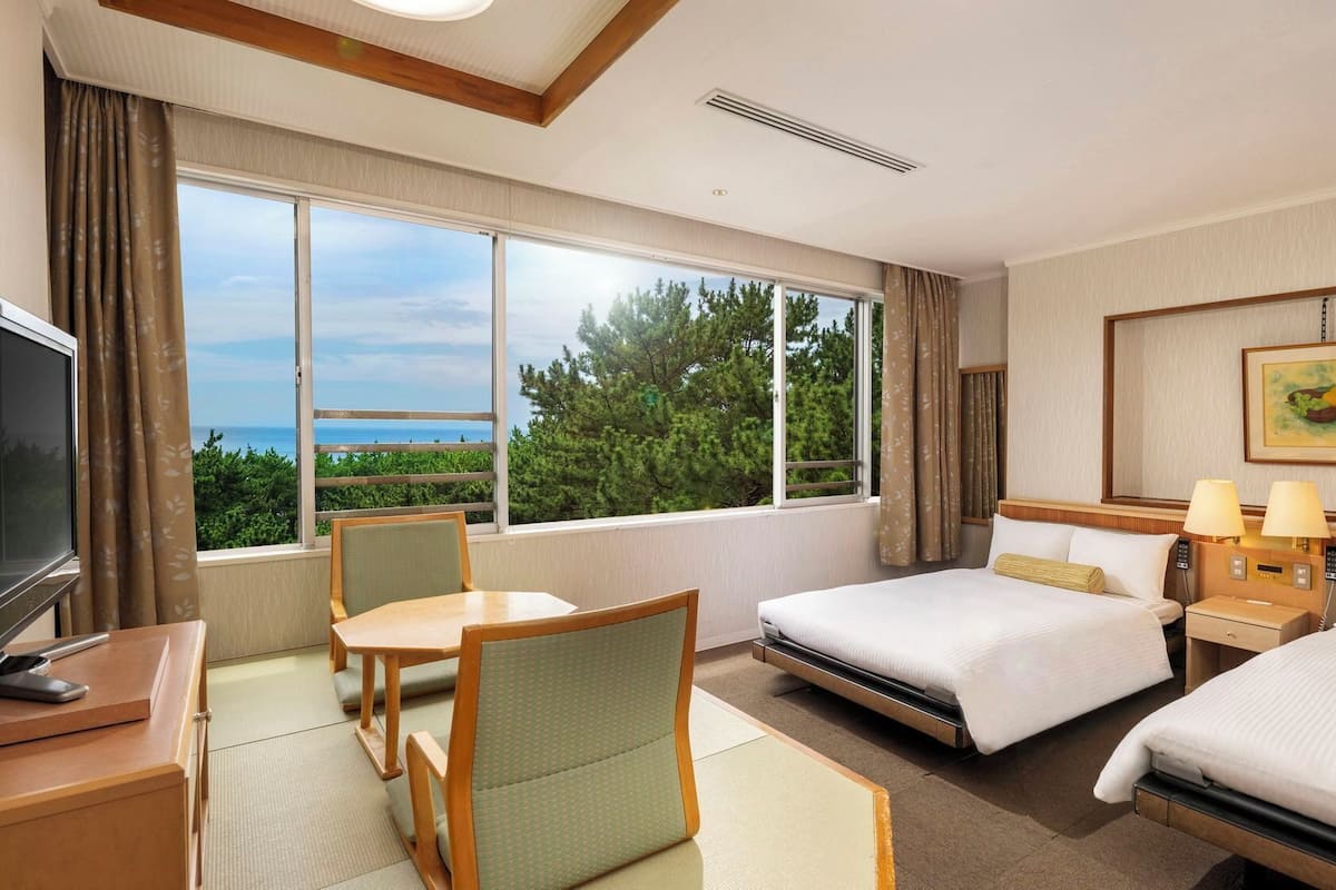 Habitación de hotel con dos camas, un escritorio, una silla, televisión y un amplio ventanal con vistas.