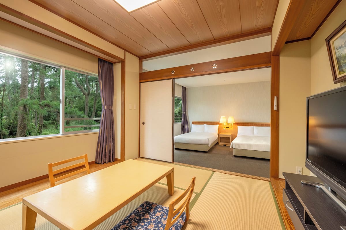 Habitación de hotel con techo de madera, un ventanal con vistas a árboles, cama con sábanas blancas, televisión y una mesita con una silla.