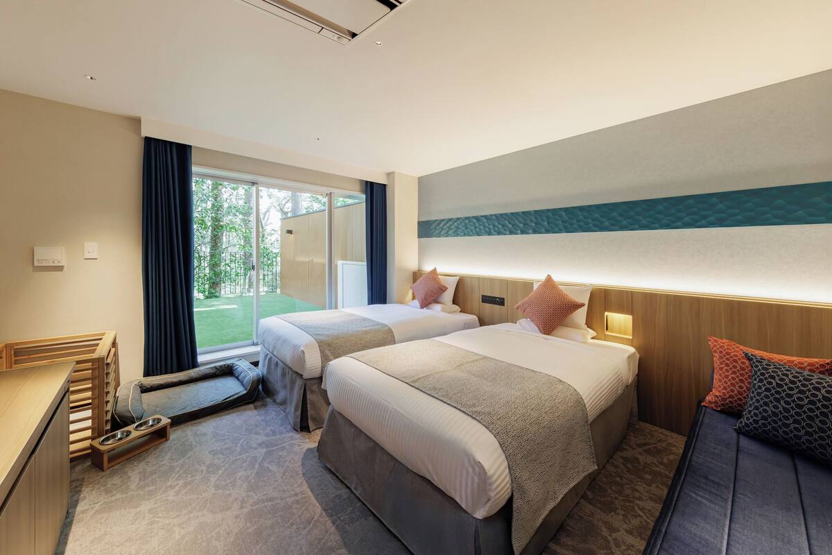 Habitación de hotel con dos camas, un escritorio con silla, una mesita con cuencos y vista al exterior.