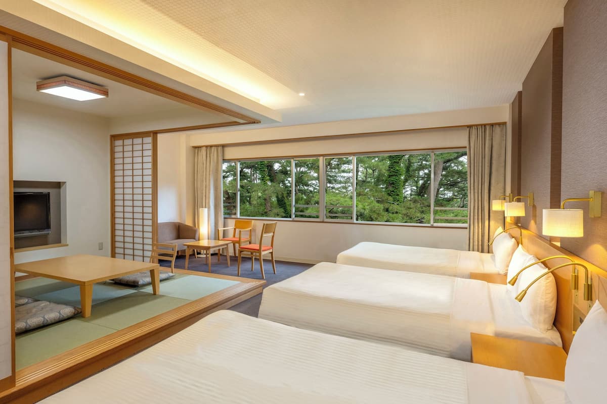 Habitación de hotel con dos camas, un escritorio, una silla, un televisor y ventanas amplias con vistas a árboles.