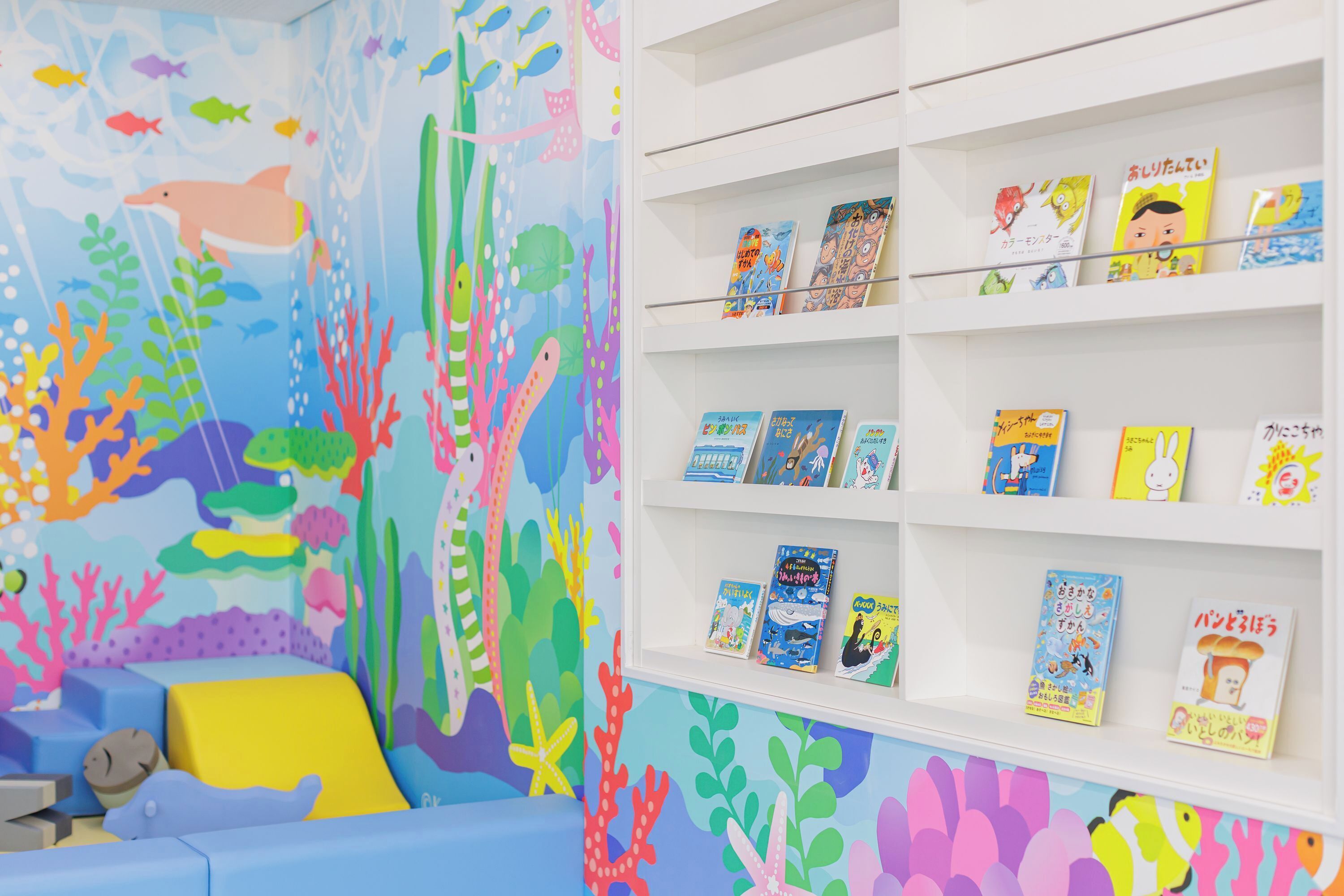 A children's room with a colorful mural of an underwater scene, a white bookshelf with children's books, and a blue cushioned seating area.