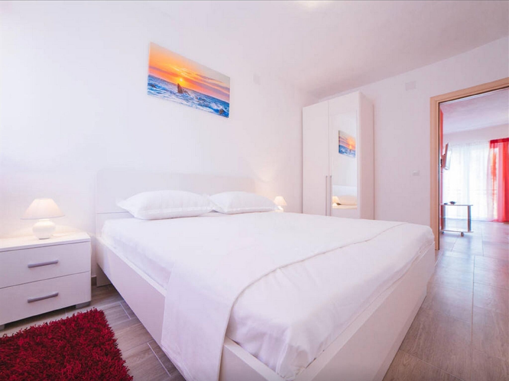 A modern bedroom with a large bed, white nightstands, a red rug, and a sunset painting on the wall.