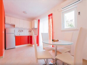 A modern kitchen with white cabinets and a red countertop, a dining area with a glass table and white chairs, and a window with red curtains.