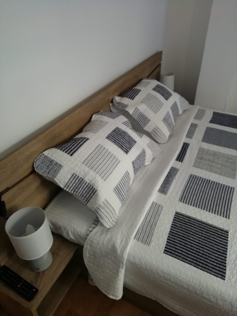 A bed with a patterned bedspread and pillows, a bedside table with a lamp and remote control, and a plain wall.