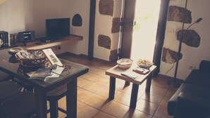 A cozy room with a tiled floor, a wooden table with a basket of snacks, a small table with a bowl of fruit, a television on a shelf, and a stone wall.