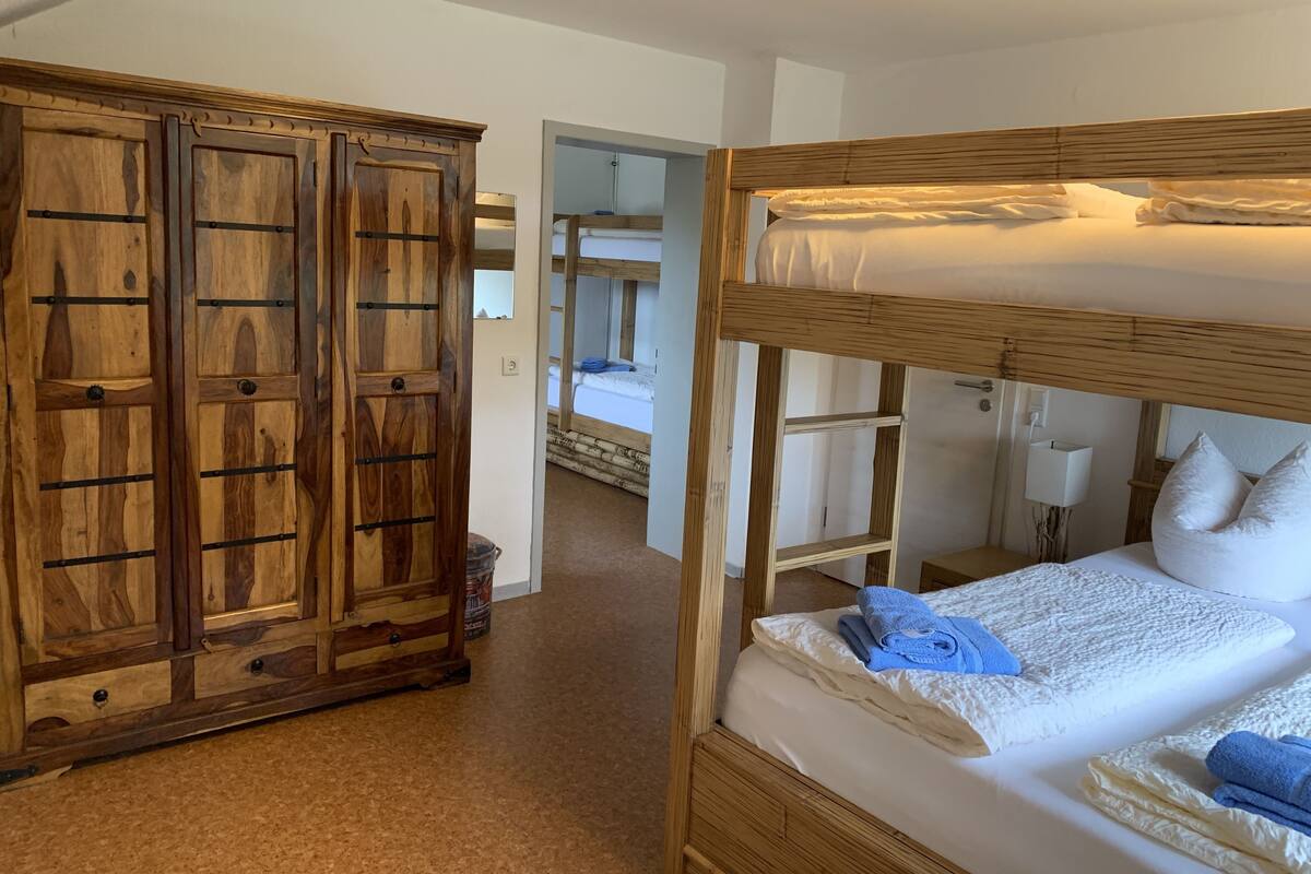 A bunk bed with white bedding and blue towels, a wooden wardrobe with multiple drawers, and a visible bathroom with a mirror and sink.