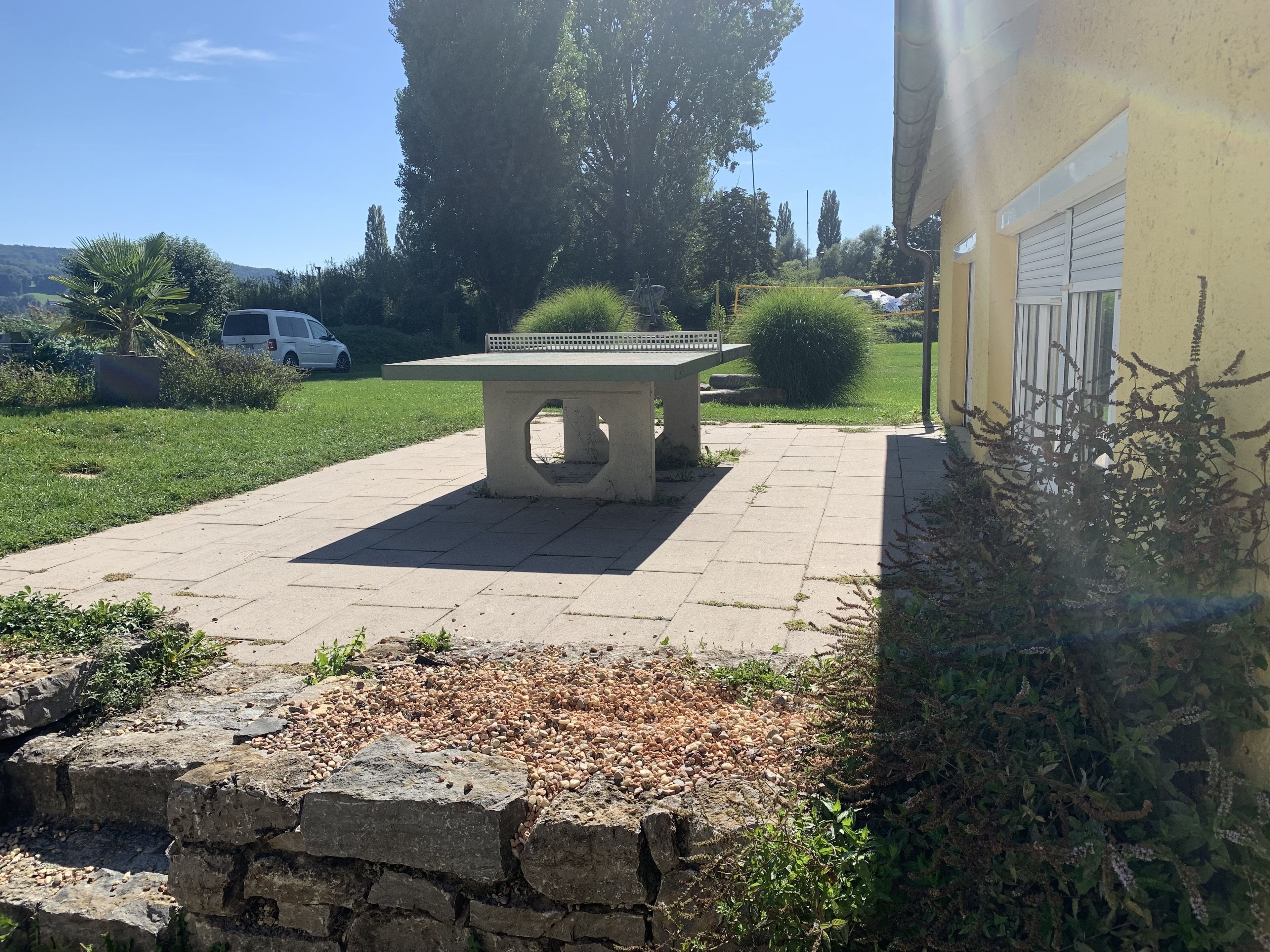 A paved patio with a stone bench, surrounded by greenery and a building with a yellow wall.