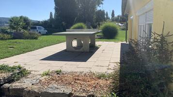 Une terrasse dallée avec un banc en pierre, entourée de verdure et bordée d’un bâtiment dont le mur est jaune.
