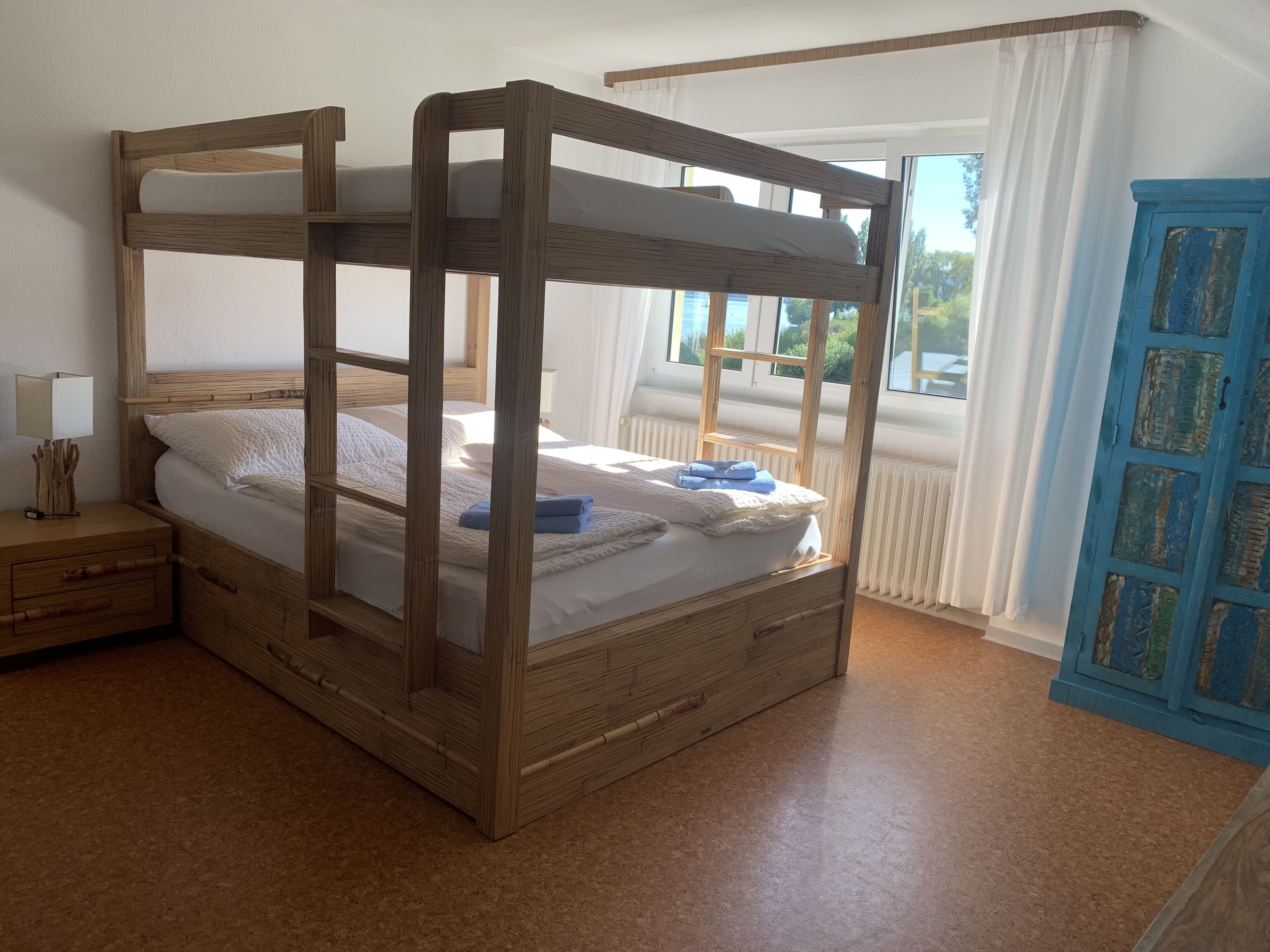 Een houten hemelbed met witte lakens en blauwe handdoeken, een nachtkastje met een lamp, en uitzicht op het landschap door een raam met witte gordijnen.