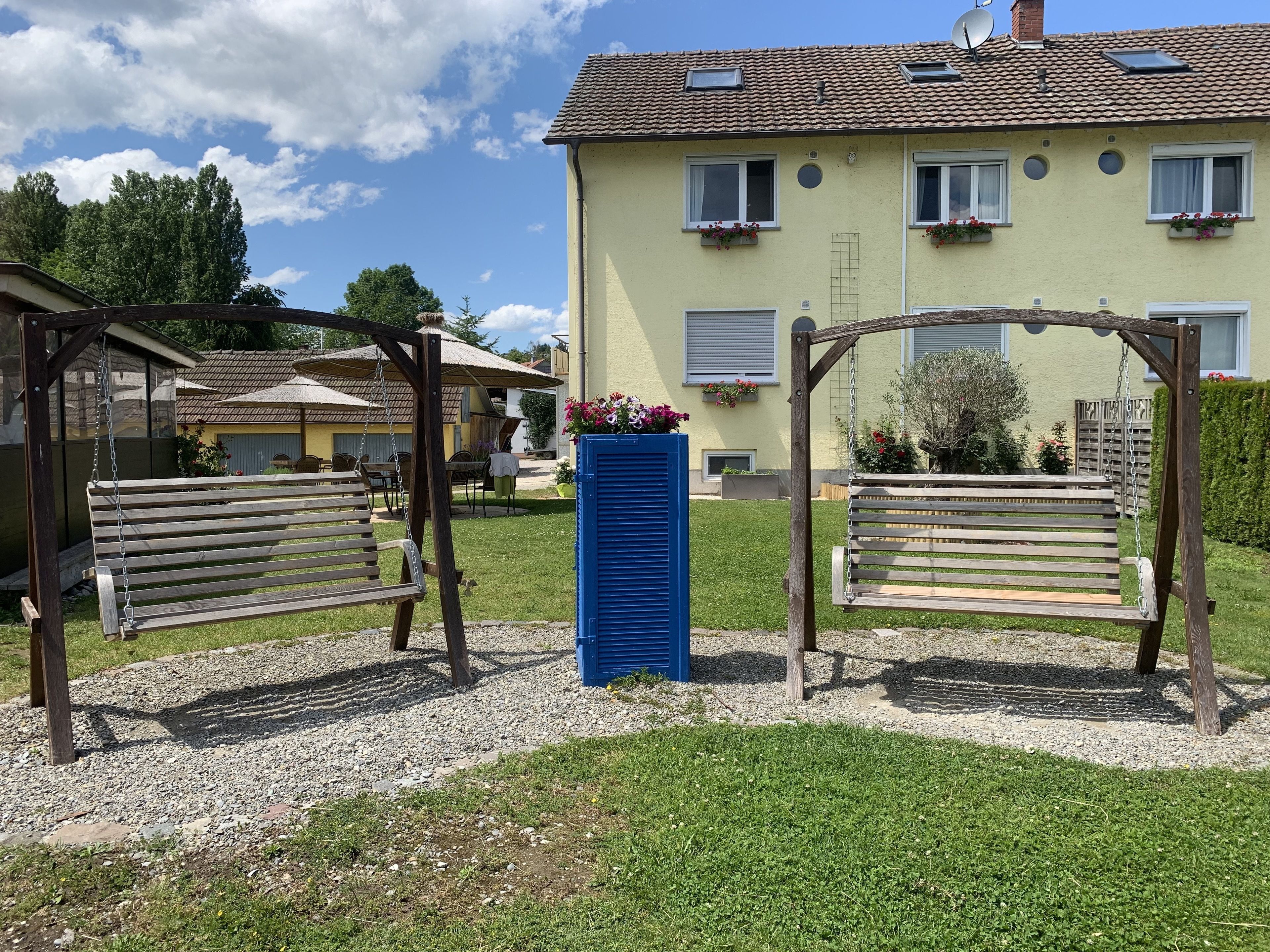 Ein Wohngebäude mit Garten, inklusive zweier Holzschaukeln und einem blauen Pflanzgefäß.