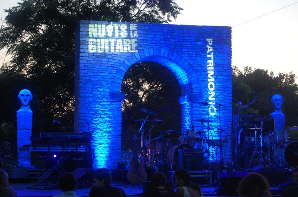 Escenario de concierto al aire libre con un arco de piedra y una banda tocando.