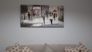 A living room with a beige sofa and patterned cushions, a painting of a street scene on the wall, and a plain white wall. - Margaritas house (Athens)