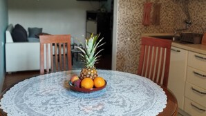 Ein Esstisch mit einer weißen Tischdecke aus Spitze, ein Holzstuhl und eine Obstschale mit einer Ananas, Orangen und Pfirsichen.
