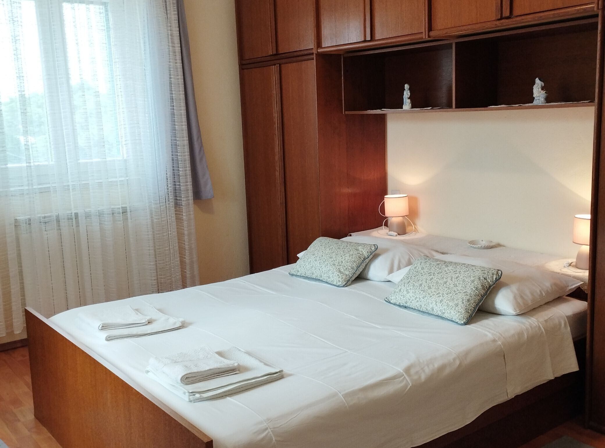 Cama bem arrumada com lençóis brancos e duas almofadas, cabeceira de madeira e mesa de cabeceira com abajur.