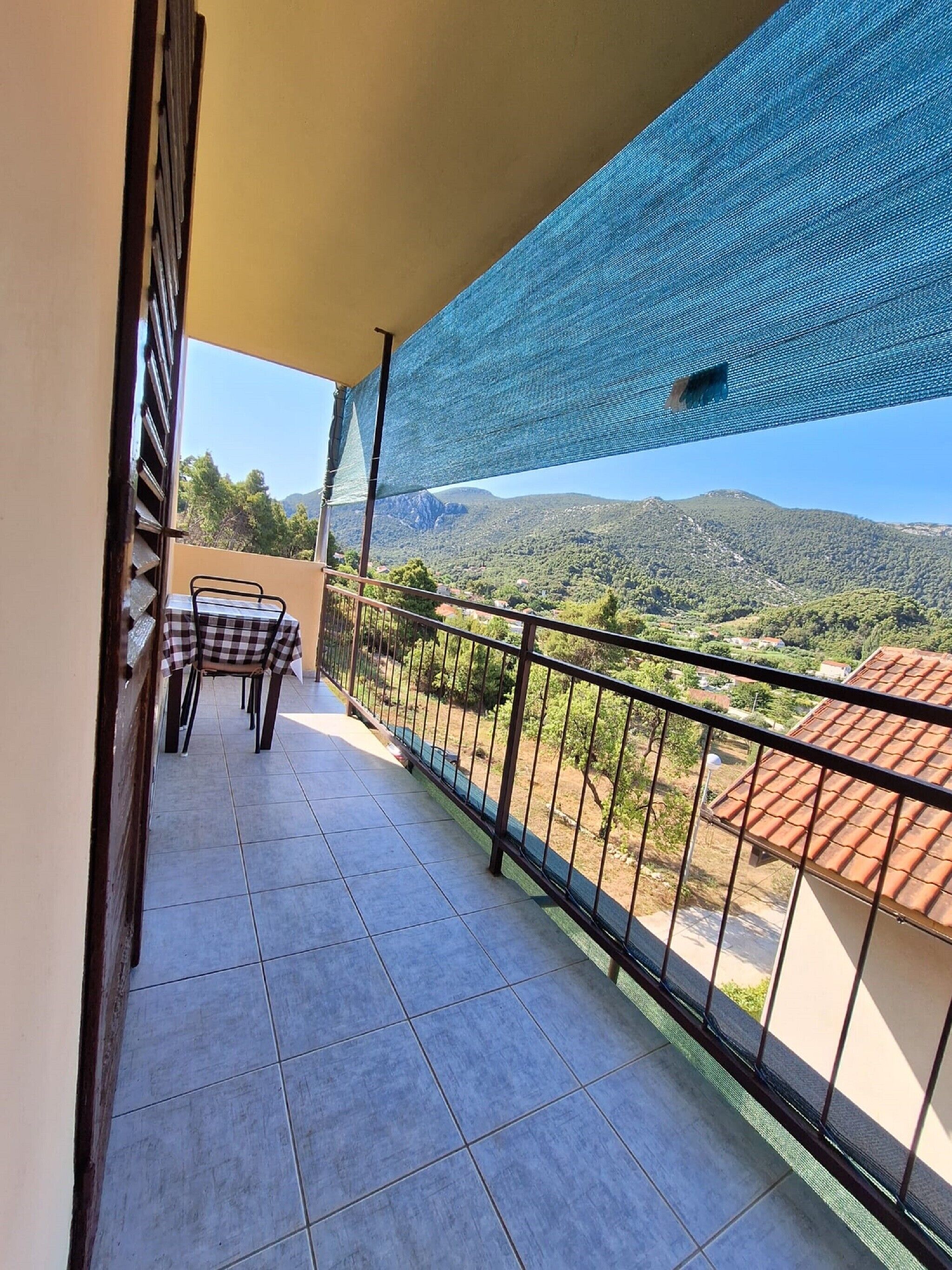 A balcony with a checkered table and chairs, a view of a mountainous landscape, and a blue mesh awning.