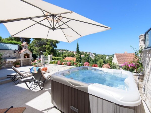 A hot tub with an umbrella, lounge chairs, and a stone fireplace in a garden setting.