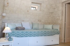 A bedroom with a stone wall, a window, a bed with patterned bedding, a bedside lamp, and a small decorative item on the nightstand.
