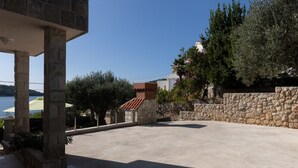 Edificio de piedra con techo de tejas rojas, zona pavimentada y vistas al mar.