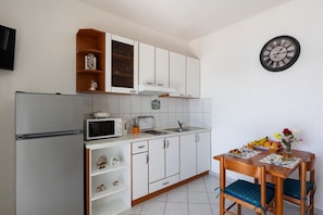 Une cuisine compacte avec des meubles blancs, un four à micro-ondes, un grille-pain et un évier. On y trouve une table à manger avec des chaises et une horloge au mur.