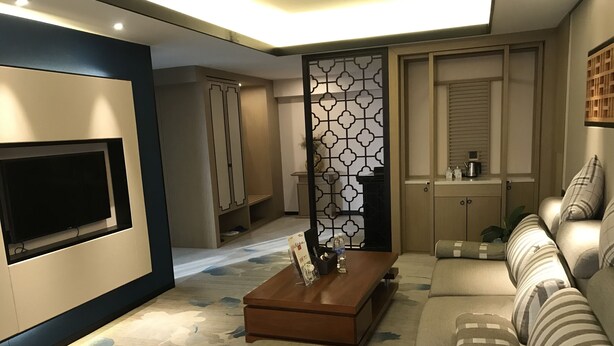A modern living room with a flat-screen TV, a wooden coffee table, a sofa with striped cushions, a decorative screen, and a cabinet with a kettle. - Metropolis Golf Hot Spring Hotel (Kunming)