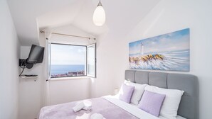 Ein Schlafzimmer mit einem Bett, einem Meeresleuchtturm Gemälde, einem Fernseher und einem Fenster mit Meerblick.