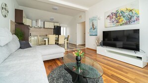 Ein modernes Wohnzimmer mit einer Couch, einem Glastisch mit Blumen, einem Flachbildfernseher und einer Küche im Hintergrund.