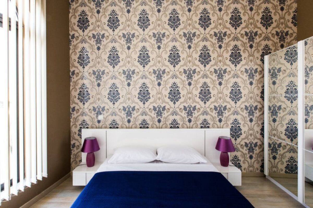 A bedroom with a patterned wallpaper, a white bed, two bedside lamps, and a wooden floor.