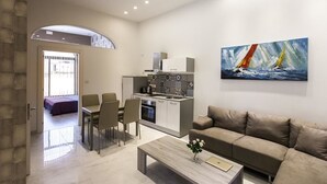 A modern living room with a sofa, a coffee table, and a dining area with chairs and a table. There is a kitchen area with a stove and cabinets. A painting of sailboats is on the wall. - Cittadella Boutique Living (Victoria)