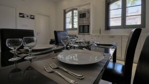 A dining area with a table set for four, including wine glasses, plates, and cutlery. There are two ovens in the kitchen area, and the room has a window with a view of buildings. - Cittadella Boutique Living (Victoria)